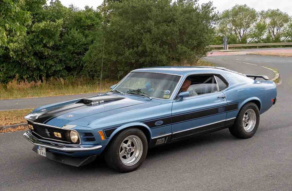 Classic 1970 Ford Mustang Mach 1 in metallic blue with black racing stripes, driving on a curved road.