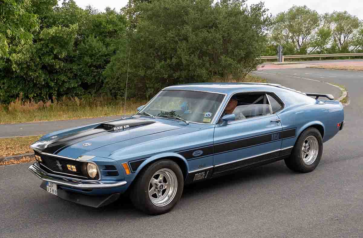 Classic 1970 Ford Mustang Mach 1 in metallic blue with black racing stripes, driving on a curved road.