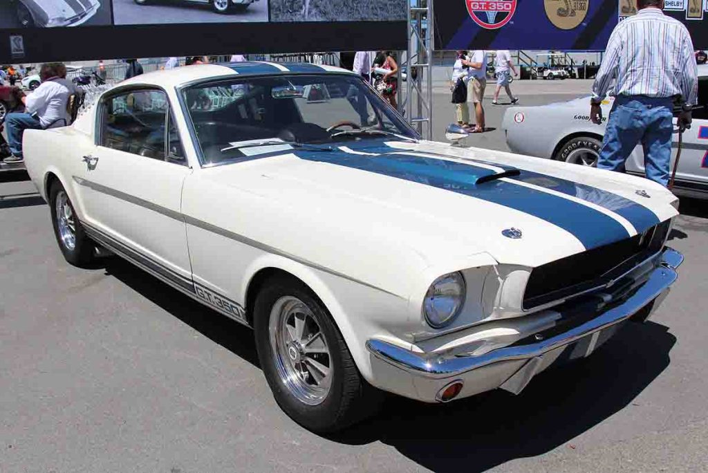 1965 Shelby Mustang GT350 in Wimbledon White with blue racing stripes parked at a vintage car show